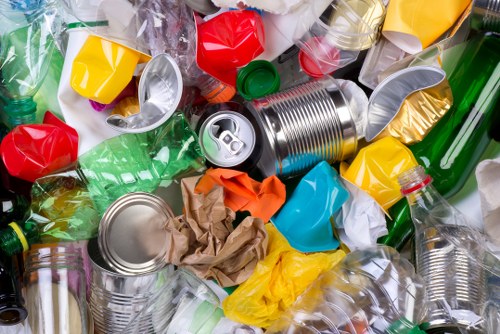 A detailed close-up image of a pile of mixed recyclable waste materials including crumpled paper, plastic bottles, aluminum cans, and plastic containers of various colors such as green, red, yellow, and clear. The waste is scattered on a flat surface, with some cans and bottles partially crushed or dented, showing textures like shiny metal and smooth plastic. The scene appears well-lit, highlighting the reflective surfaces of the aluminum cans and the vibrant colors of the plastic items, which are typical of materials requiring disposal and collection by commercial waste removal services. This image exemplifies the type of waste collections handled by Commercial Waste Removal Westminster in the Westminster area, illustrating the importance of segregating recyclable rubbish and maintaining clean premises. The background is indistinct, focusing attention solely on the cluttered mixture of trash in the foreground, conducive to environmental waste management practices and local rubbish removal work.