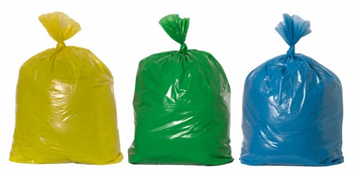 The image displays three large rubbish bags placed on a neutral surface, each made of shiny, thick plastic with a crinkled texture. The first bag on the left is bright yellow, the middle bag is vivid green, and the right bag is royal blue. All three are tightly closed at the top with the plastic gathered and knotted, indicating they are filled with waste material. The bags are positioned side by side, with the yellow bag slightly in front, the green bag to its right, and the blue bag further to the right. The background is plain and unobstructed, suggesting an outdoor or indoor setting suitable for waste collection. The overall scene reflects typical rubbish collection packaging, relevant to commercial waste removal services in Westminster and surrounding areas, emphasizing a straightforward and professional approach to waste disposal. Commercial Waste Removal Westminster often handles waste represented by such standard refuse bags, as seen here, ready for collection or disposal.
