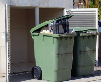 Two large green wheelie bins, one with a black lid that is open, filled with mixed waste including cardboard boxes and plastic packaging, are positioned in front of a white outdoor enclosure or shelter on a paved surface. The bins are made of durable plastic with textured surfaces and black wheels for mobility. The shelter above them offers partial cover and appears to be part of a residential or commercial waste collection area, with a beige wall and partial fencing visible in the background. The image reflects a typical scene of rubbish awaiting collection or disposal, common in waste management services around Westminster. Commercial Waste Removal Westminster operates in this locality, providing rubbish removal solutions, as suggested by the surrounding environment and street setting.