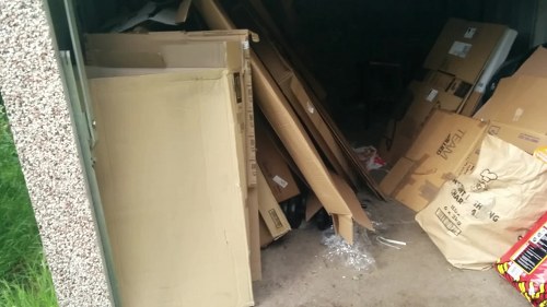 A collection of discarded items including a partially visible cream or beige piece of furniture with a textured surface leaning against a wall, surrounded by various cardboard boxes, some flattened and others partly intact, along with crumpled packaging material and clear plastic wrappers. The items are situated on a concrete surface, possibly a driveway or storage area, with a small patch of grass visible on the left side of the image. The background appears to be an interior or garage space with additional boxes resting against the back wall. The scene reflects typical rubbish awaiting collection or disposal, as seen in services like those provided by Commercial Waste Removal Westminster for commercial or domestic waste clearance in Westminster or nearby areas.