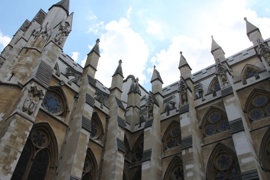 A street scene featuring historic architecture with two prominent tall, twin Gothic-style church towers constructed from light-coloured stone, adorned with intricate detailing and pointed spires, set 